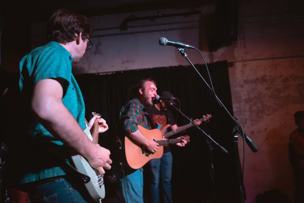 Two musicians perform on stage at an indoor venue, one singing at a microphone while the other plays electric guitar.