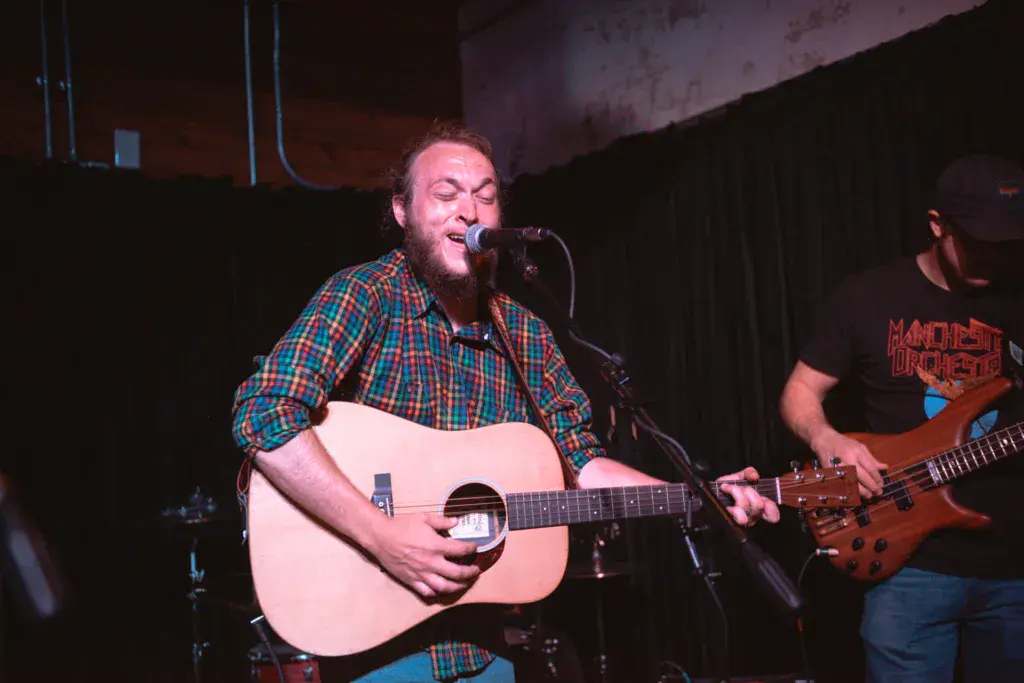 A bearded musician performs with an acoustic guitar at a microphone while a bassist plays alongside on stage.