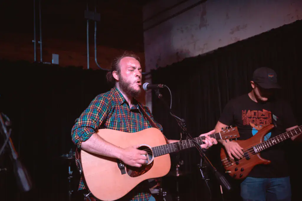 A bearded musician in a plaid shirt plays acoustic guitar at a microphone while a bassist stands nearby on stage.