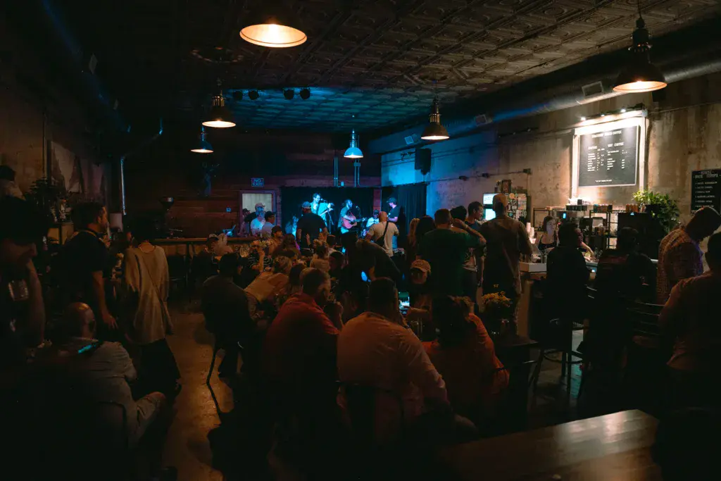 A crowd of festival attendees fills an indoor venue with blue lighting while a band performs on stage in the background.