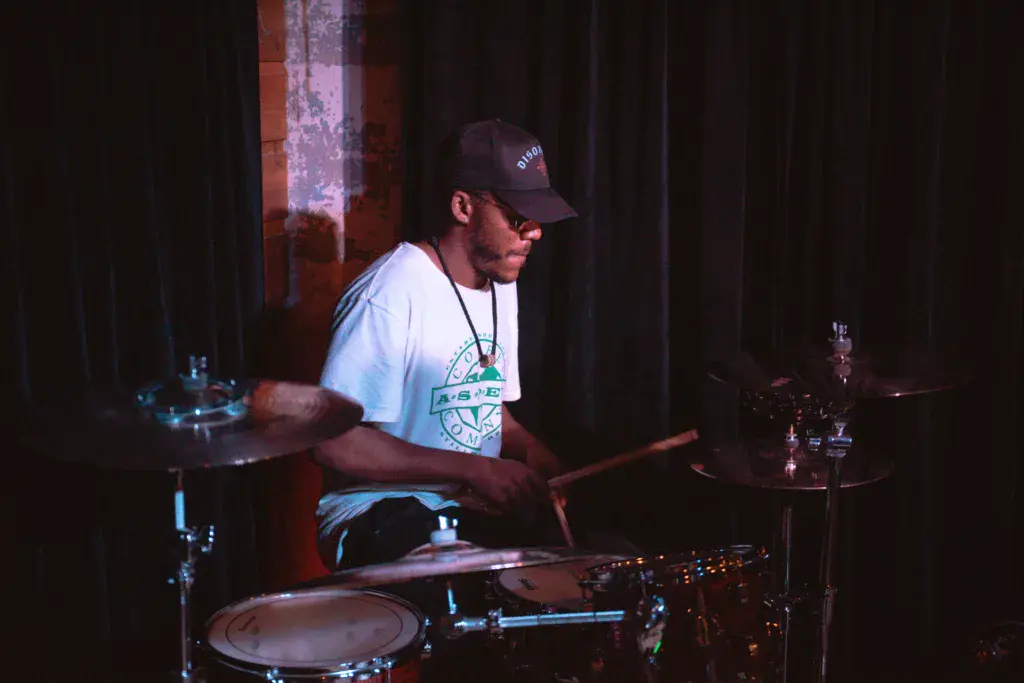 A drummer performs on stage wearing a baseball cap and light-colored t-shirt during an evening performance.