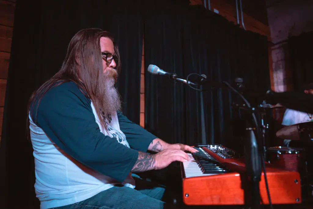 A performer with long beard and glasses plays a red keyboard synthesizer on stage at Dancing Rabbit Music Festival.