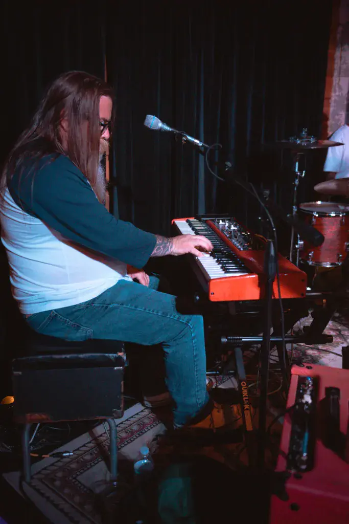 A performer with long dark hair plays a red keyboard synthesizer on stage during an evening performance.