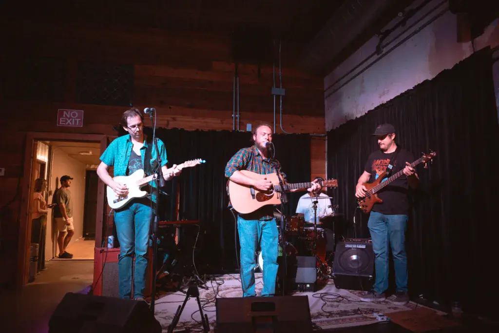 A live band performs on stage with electric and acoustic guitars and bass, with a drummer visible in the background.