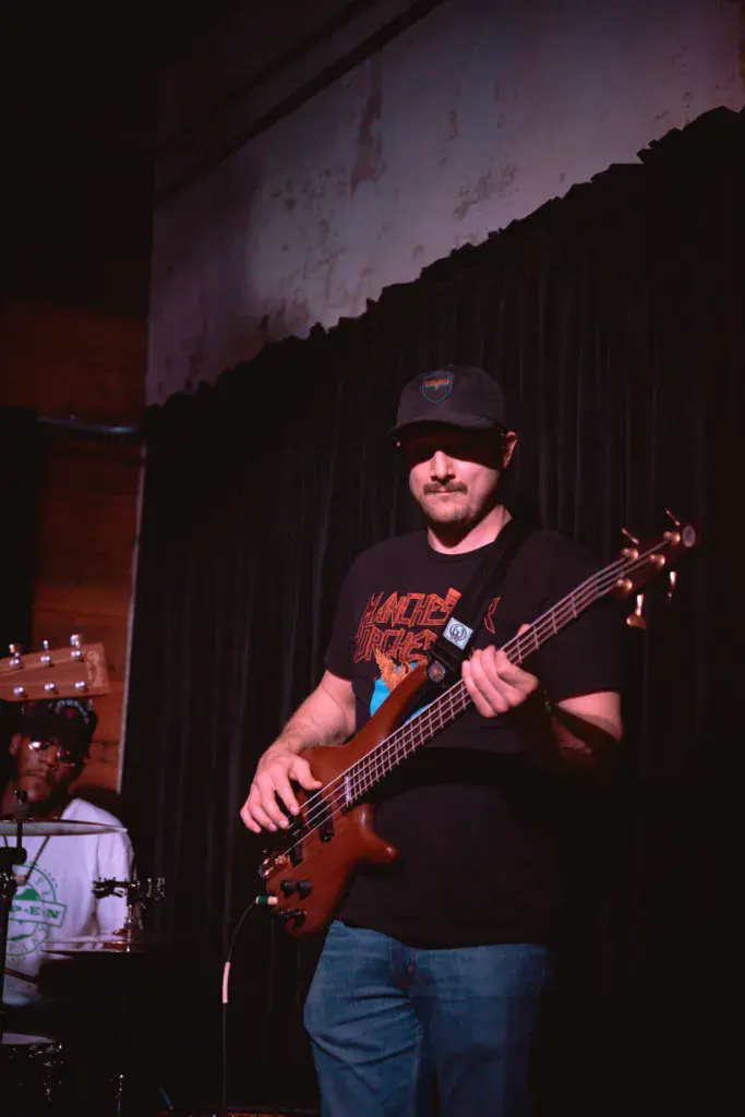 A bass guitarist performs on stage wearing a black cap and graphic t-shirt, playing a red bass guitar.