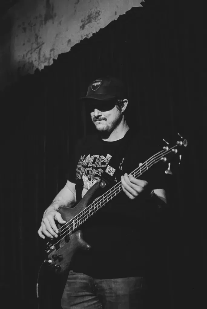 A bass guitarist performs on stage wearing a black t-shirt, cap, and sunglasses against a dark backdrop.