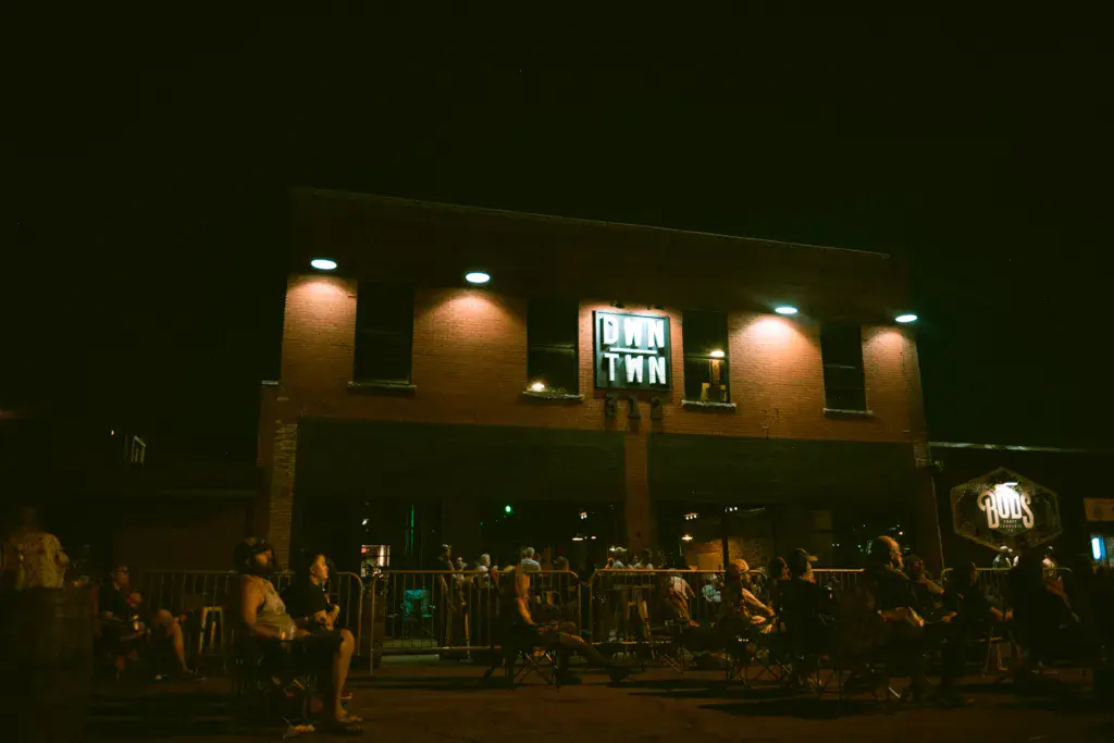 Nighttime venue with crowd gathered behind barriers in front of an illuminated brick building with neon signage.