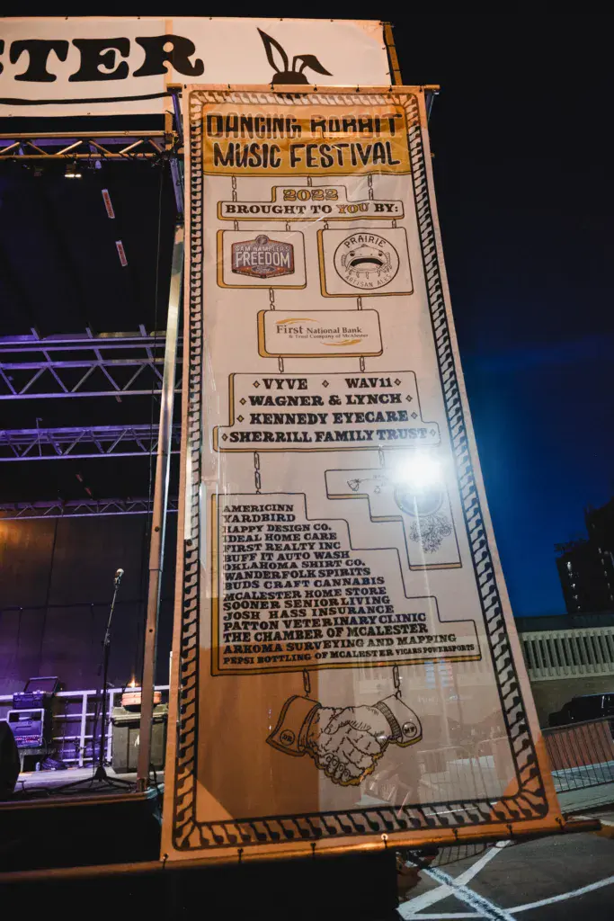 Large wooden signage for Dancing Rabbit Music Festival displaying sponsor logos and event information at night.