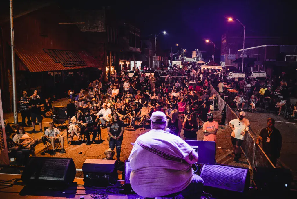 A performer on stage addresses a large crowd gathered in an outdoor street venue at night.