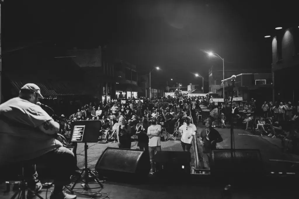 A musician performs on stage at night while a large crowd gathers in an urban venue under street lights.