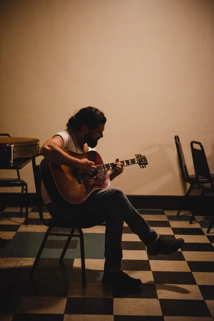 A performer sits on a chair playing an acoustic guitar on a checkered floor stage.