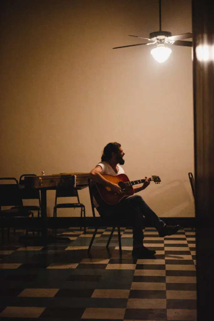 A bearded musician sitting alone on a chair, playing an acoustic guitar in an indoor venue under warm ceiling lighting.