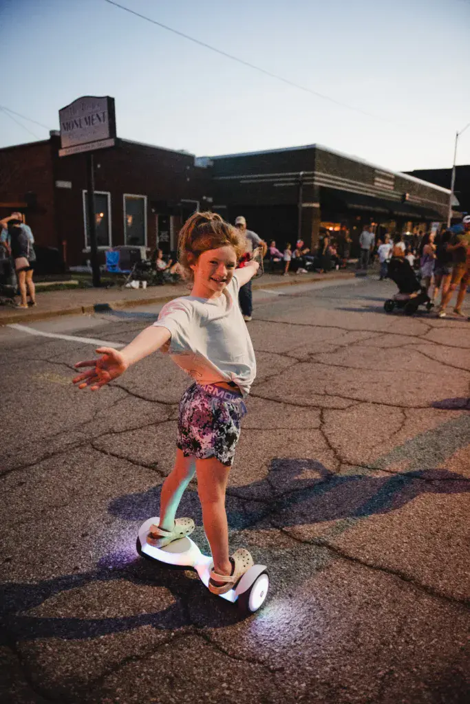A young child rides a hoverboard while smiling at the camera in front of a venue building at the festival.
