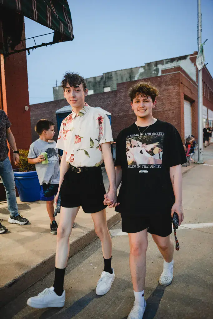 Two young people hold hands while walking through an outdoor festival venue with brick buildings in the background.
