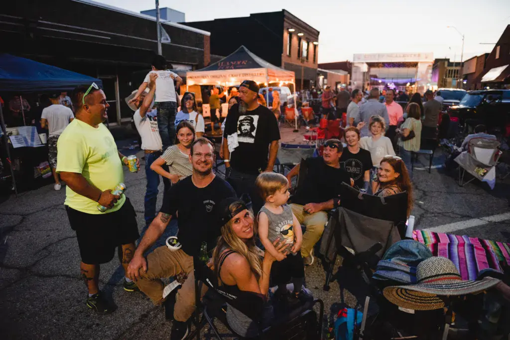 A diverse crowd of festival-goers gathers in an urban outdoor venue with vendor tents and buildings in the background.
