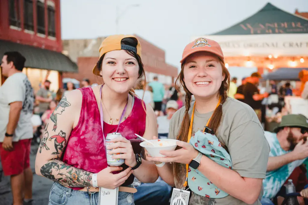 Two smiling festival attendees holding food and drinks at an outdoor event with crowds and buildings in the background.