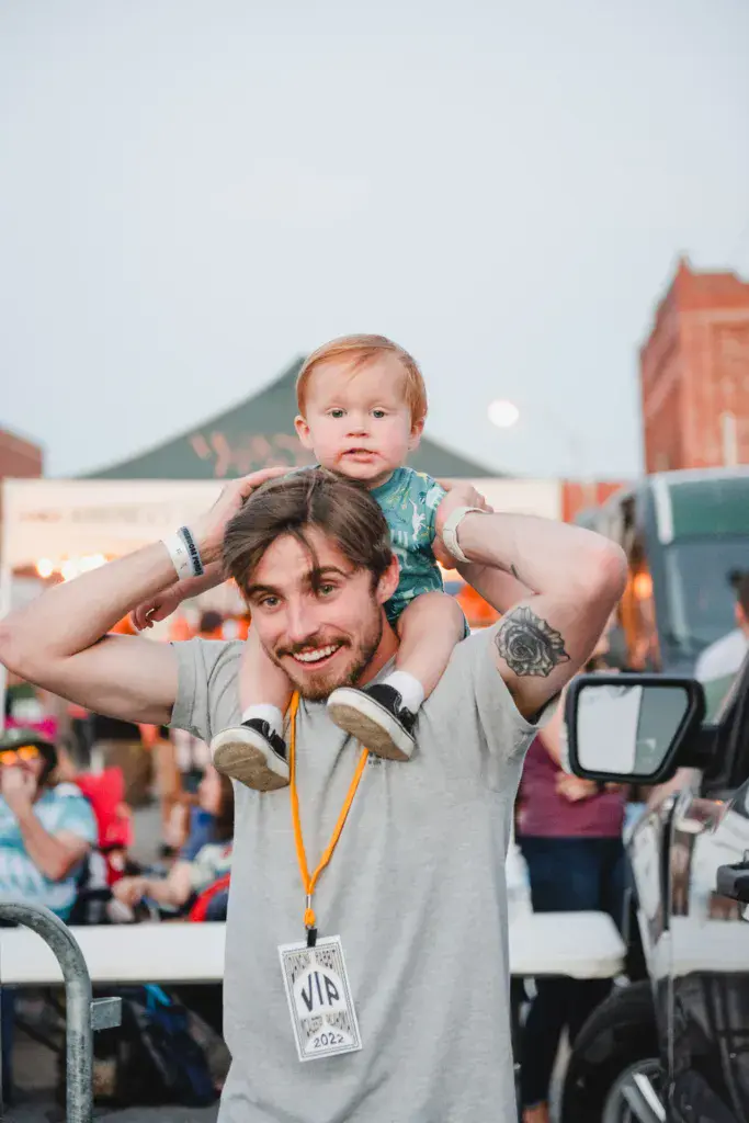 A man wearing a VIP lanyard holds a small child on his shoulders at an outdoor music festival with crowds and venue structures visible behind them.