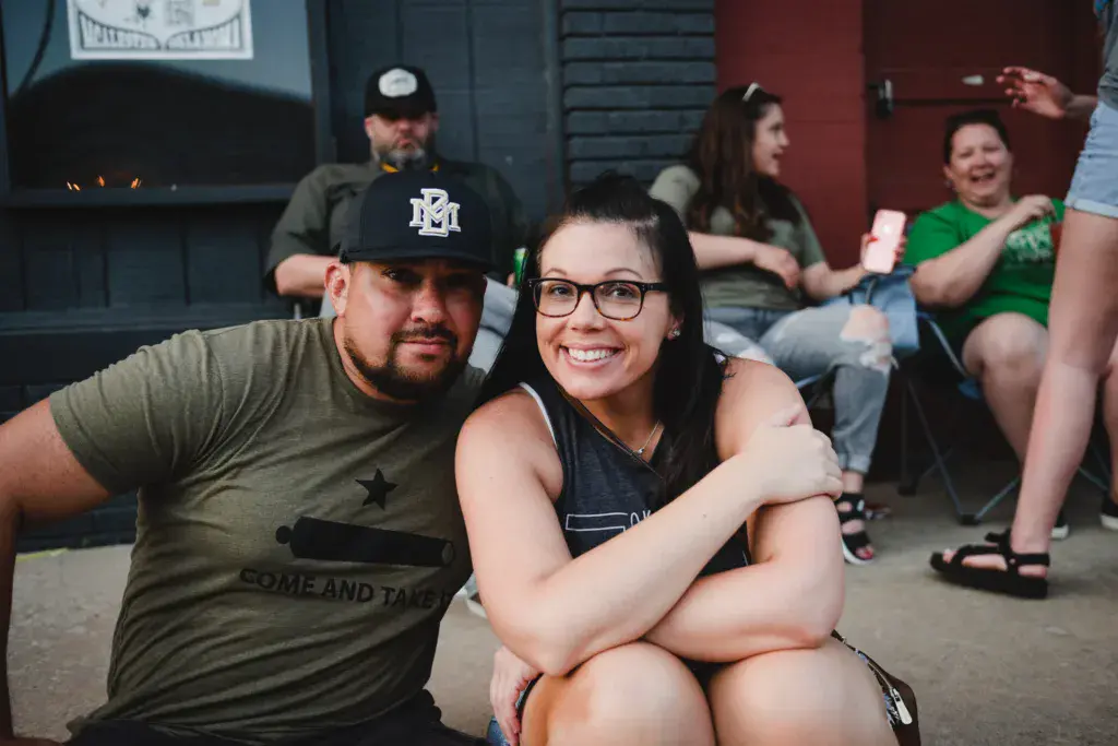 Two smiling attendees pose together at an outdoor venue with other festival-goers visible in the background.