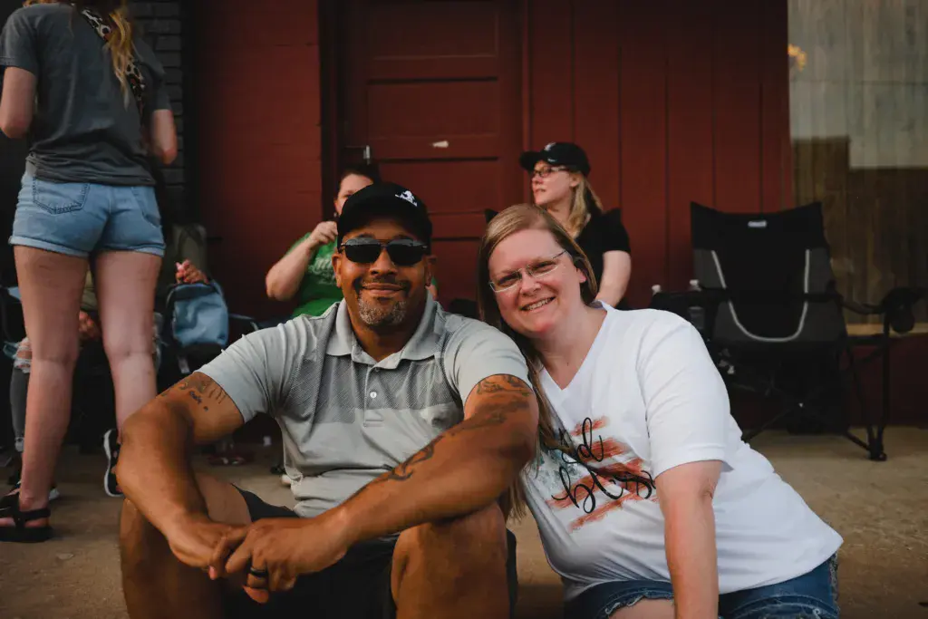 Two people smile at the camera while seated outdoors at an event, with others visible in the background.