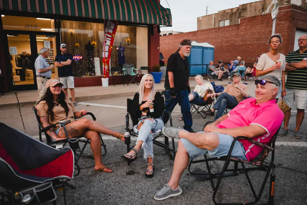 Festival attendees relax in folding chairs in a parking lot venue with brick buildings and storefronts nearby.