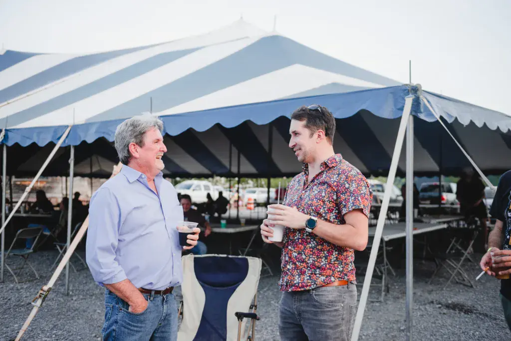 Two men holding drinks chat and smile under a large striped blue and white tent at an outdoor festival.