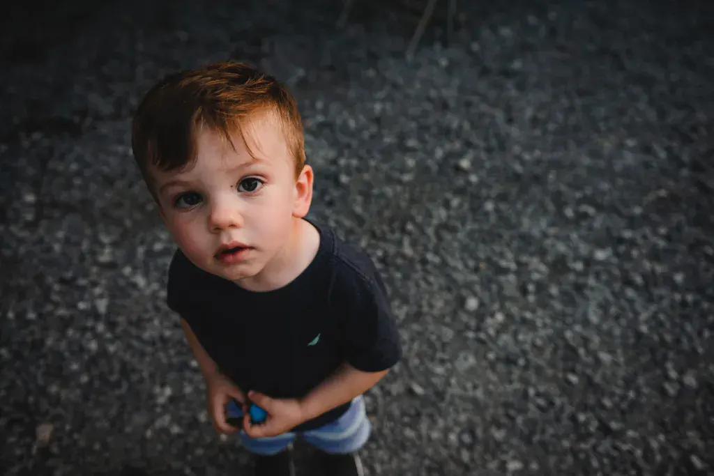 A young child with reddish-brown hair stands on a gravel surface looking up at the camera.