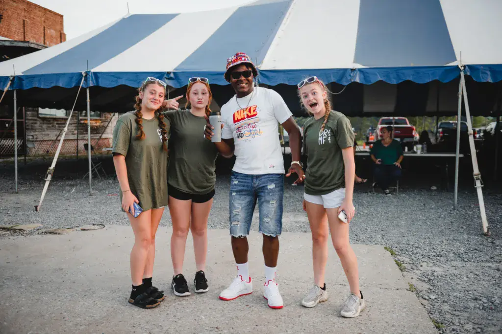 Four people pose together for a group photo under a blue and white striped tent at an outdoor festival.