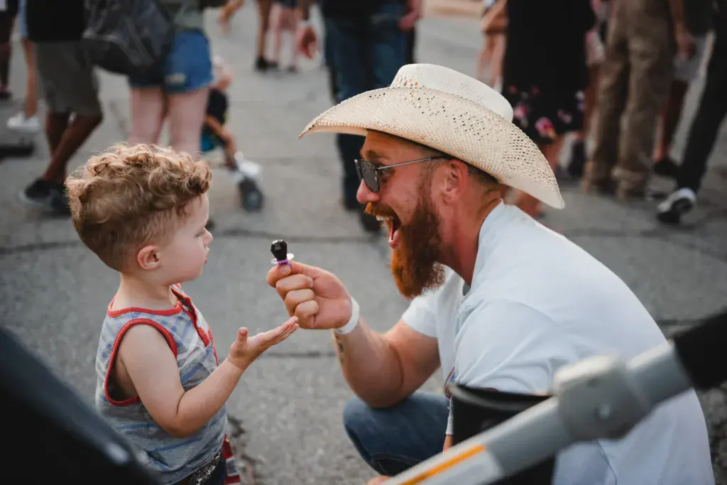 A man in a cowboy hat playfully interacts with a young child at a festival venue.