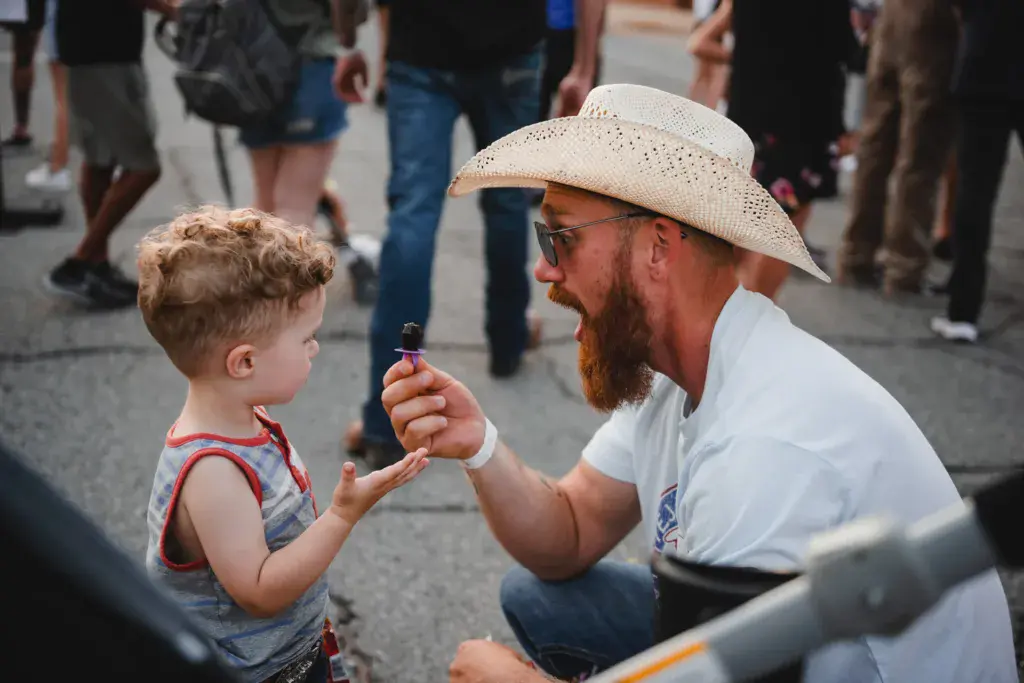 A man in a cowboy hat hands a small item to a young child at a festival gathering.