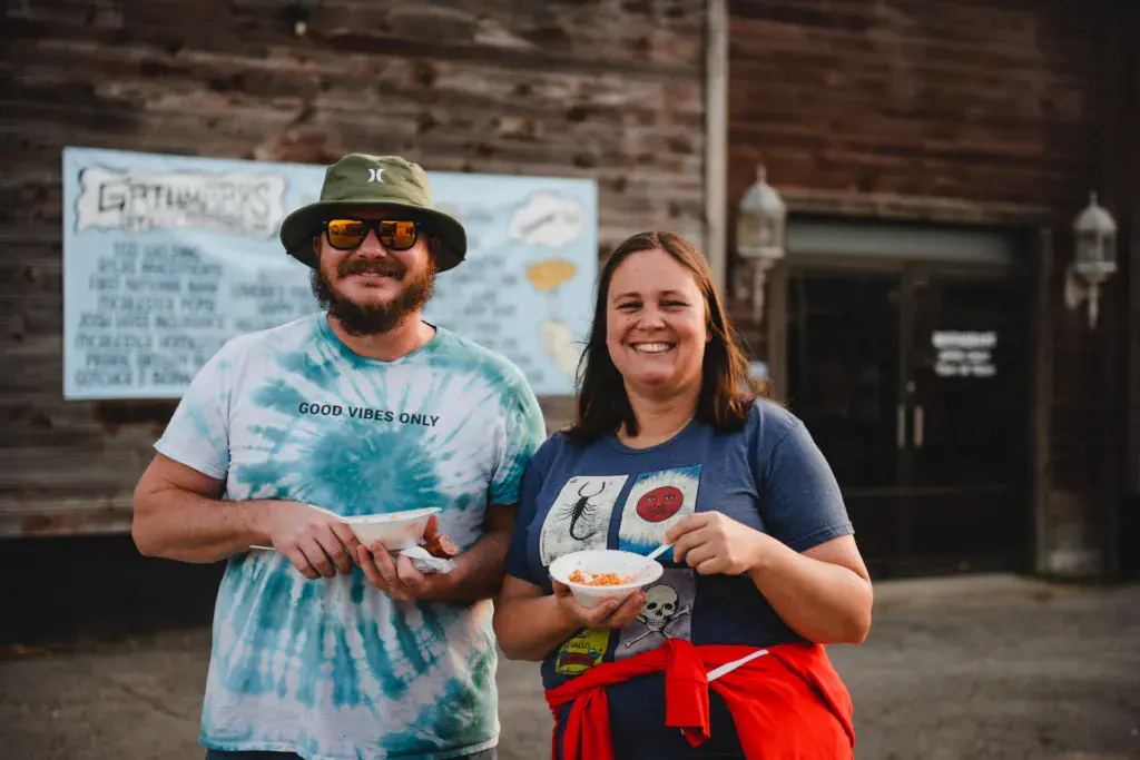 Two people smiling while holding food at an outdoor festival venue with a wooden building backdrop.