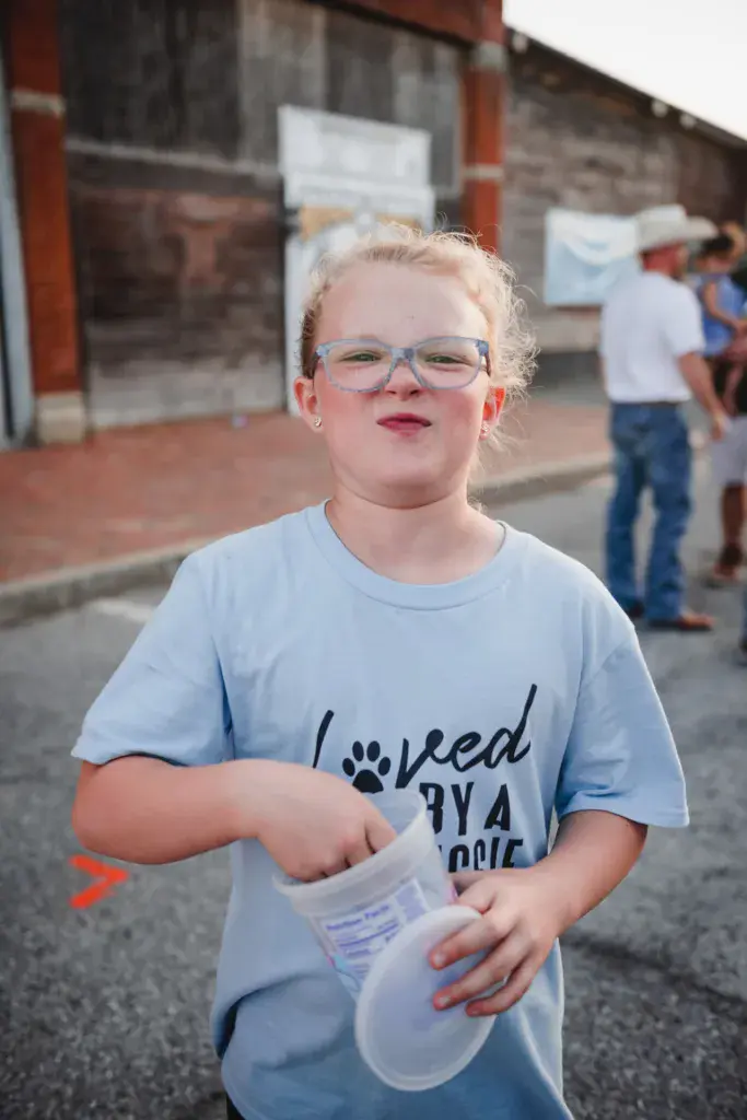 Young child wearing blue glasses and a light blue t-shirt smiles at the camera while holding a cup at an outdoor venue.