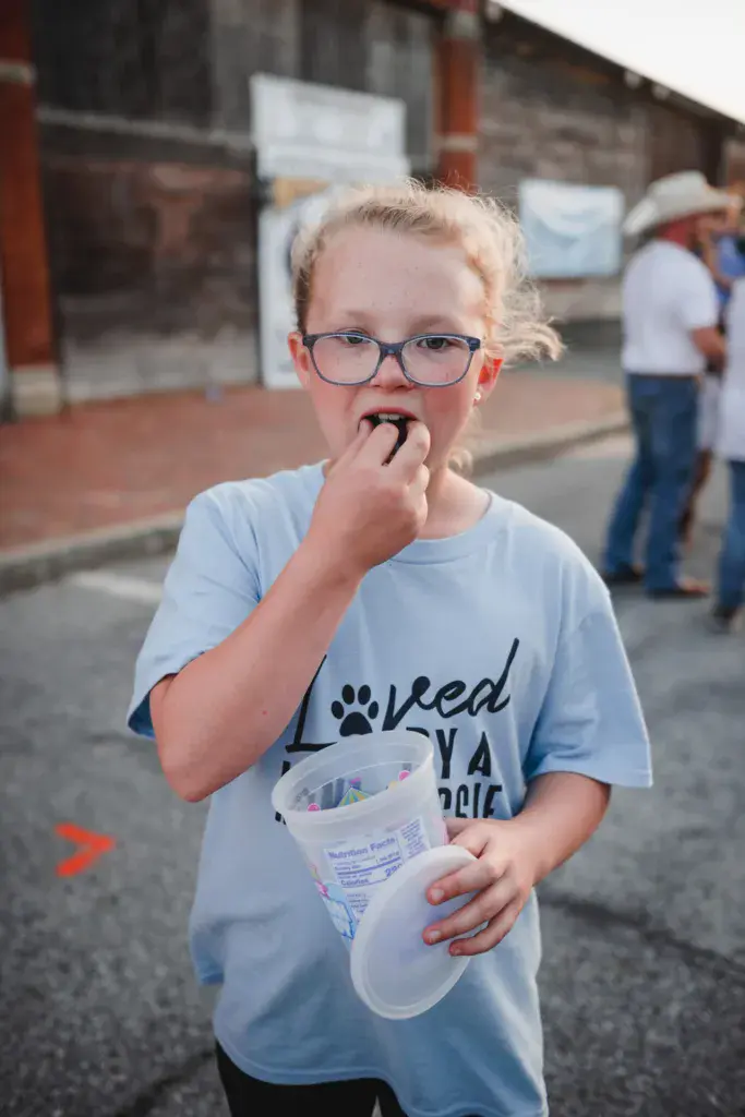Young child wearing glasses and a blue shirt eating from a cup at an outdoor festival venue.