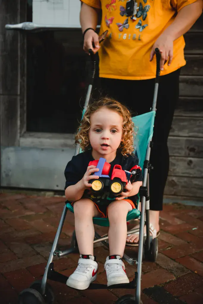 Young child in a stroller holds a toy truck while an adult wearing a yellow graphic shirt stands behind.