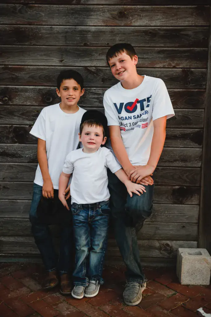Three young boys smiling at the camera while posing together against a rustic wooden wall.