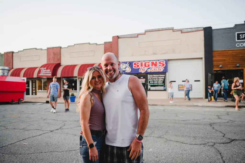 Two people smile for the camera at an outdoor festival venue with storefronts and festival-goers in the background.