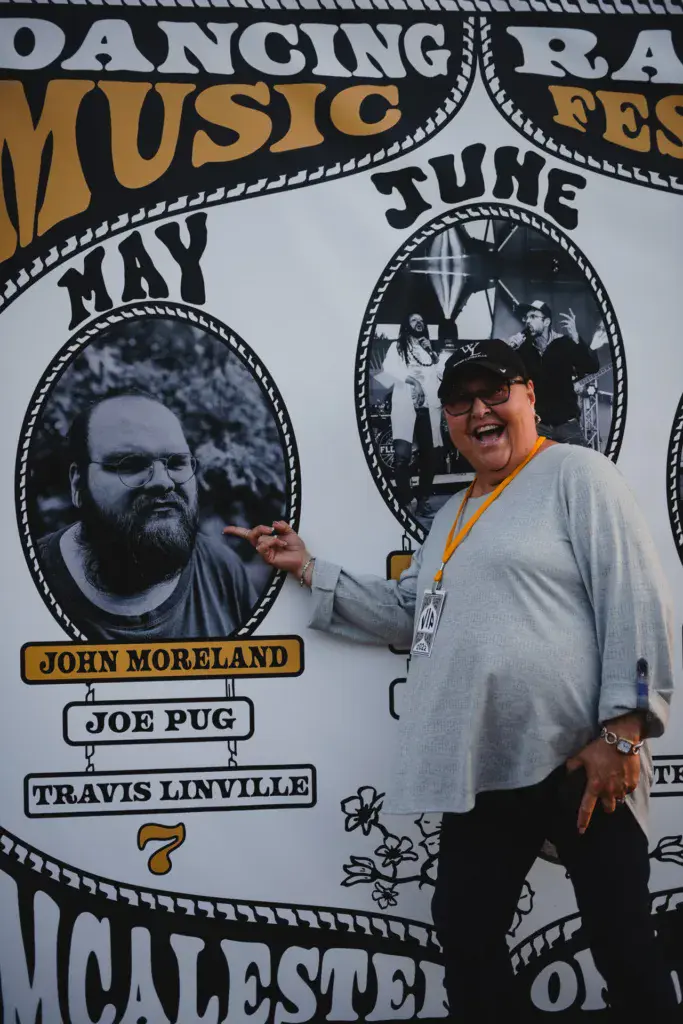 Festival attendee posing in front of a large banner displaying performer portraits and names at Dancing Rabbit Music Festival.