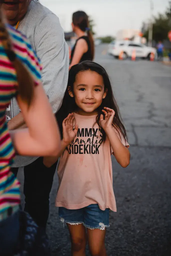 A young child smiles at the camera while standing with adults in a festival parking lot.