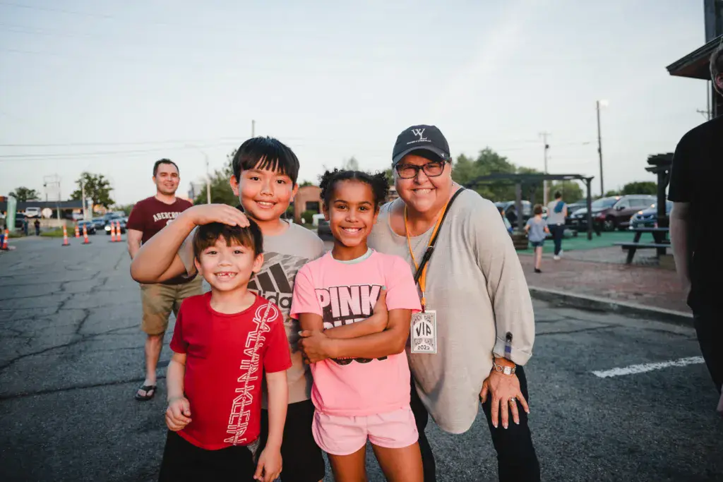 A group of adults and children posing together at an outdoor festival venue with parking lot and structures visible in the background.
