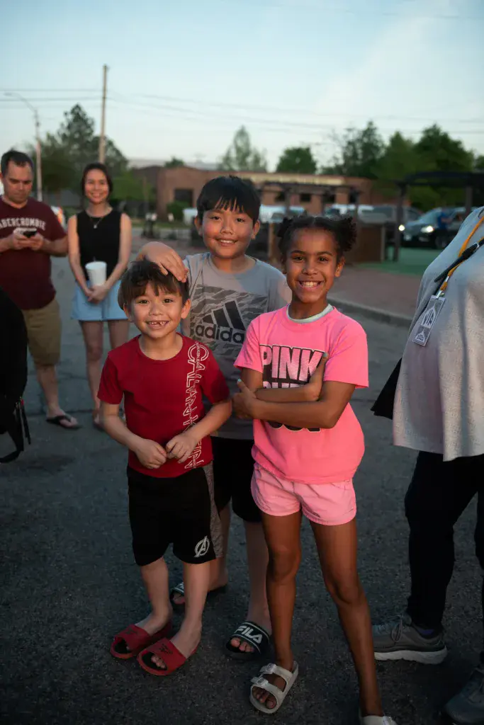 Children smiling at the camera while standing in a parking lot at an outdoor music festival.