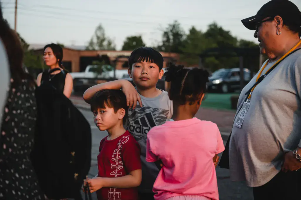 Children and adults gather in a casual group in an outdoor venue parking area with trees and buildings visible in the background.