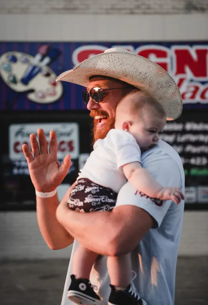 A man in a cowboy hat and sunglasses holds a small child while waving at a festival venue with sponsor signage visible behind them.