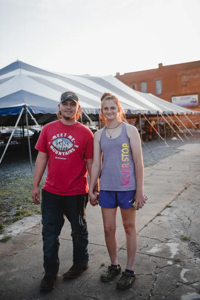 Two young attendees pose together at the Dancing Rabbit Music Festival venue with white tents and a brick building behind them.