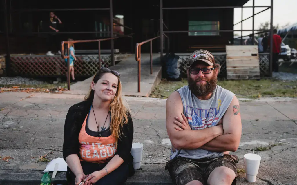 Two attendees sit together on the ground at the festival venue with the stage structure visible behind them.