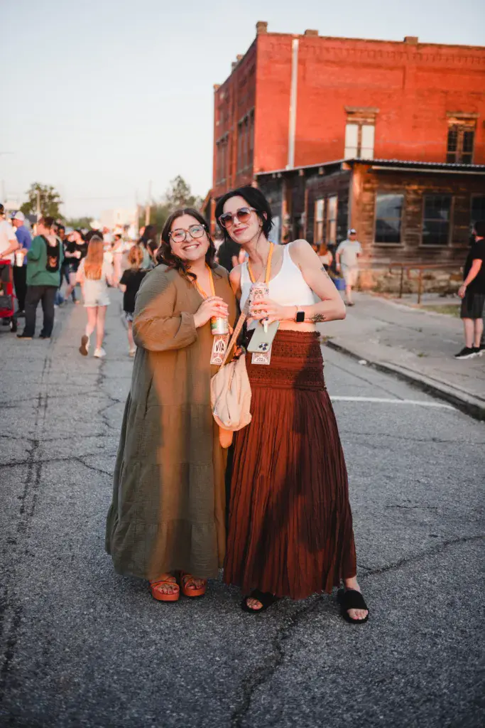 Two festival attendees with lanyards smile at the camera on a street in front of a historic red brick building.