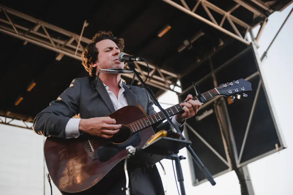 A performer sings into a microphone while playing an acoustic guitar on an outdoor festival stage.