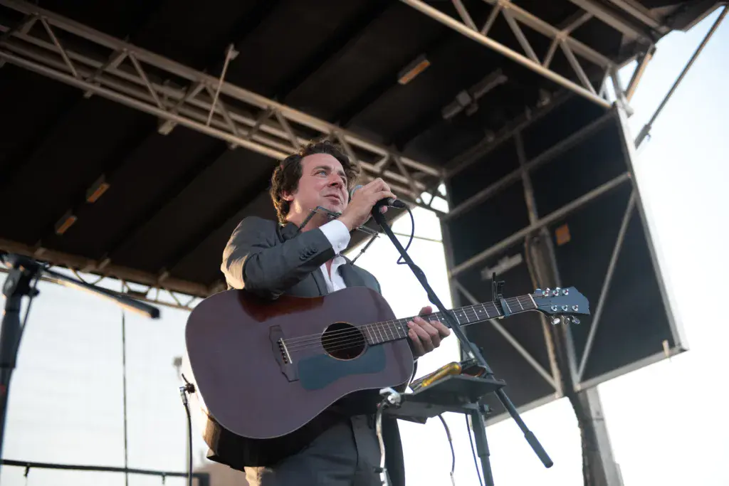 A performer on stage plays an acoustic guitar and sings into a microphone under a metal roof structure.