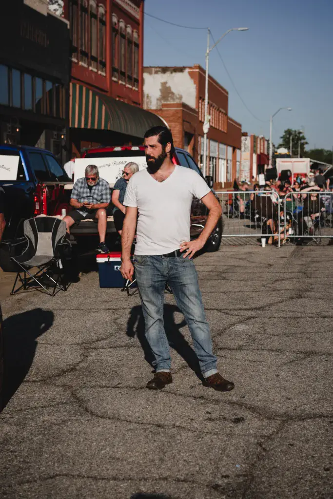 A bearded man in a white t-shirt stands in a parking lot at an outdoor music festival with vintage brick buildings and spectators in the background.