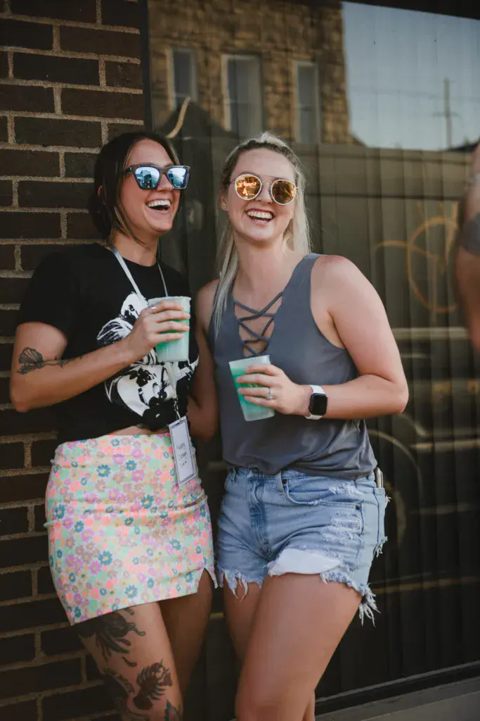 Two smiling festival attendees wearing sunglasses and holding drinks stand against a brick building.
