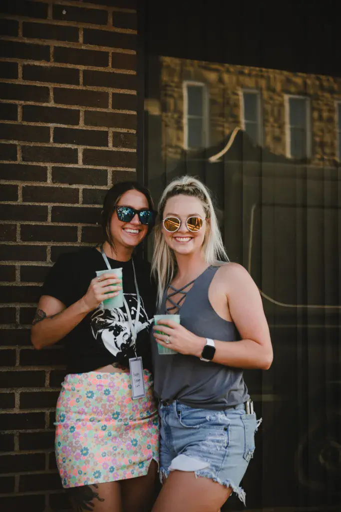 Two festival attendees in sunglasses smile while holding drinks against a brick building at the Dancing Rabbit Music Festival.