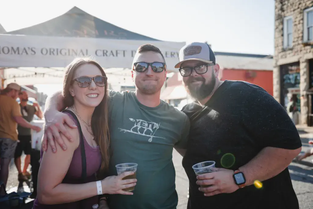 Three people smiling at the camera while holding drinks at an outdoor festival with sponsor tents visible behind them.